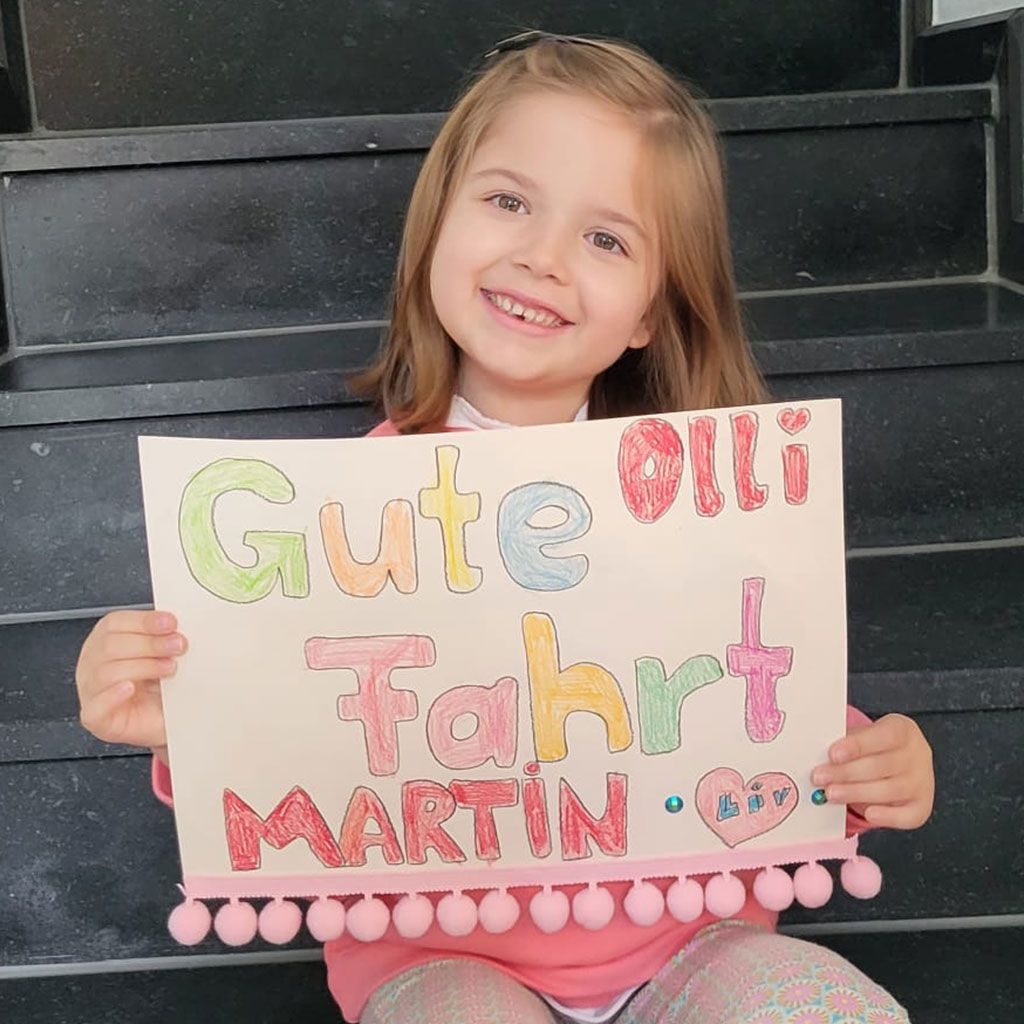 Liv hält ein selbstgemaltes Plakat mit der Aufschrift Gute Fahrt Olli und Martin